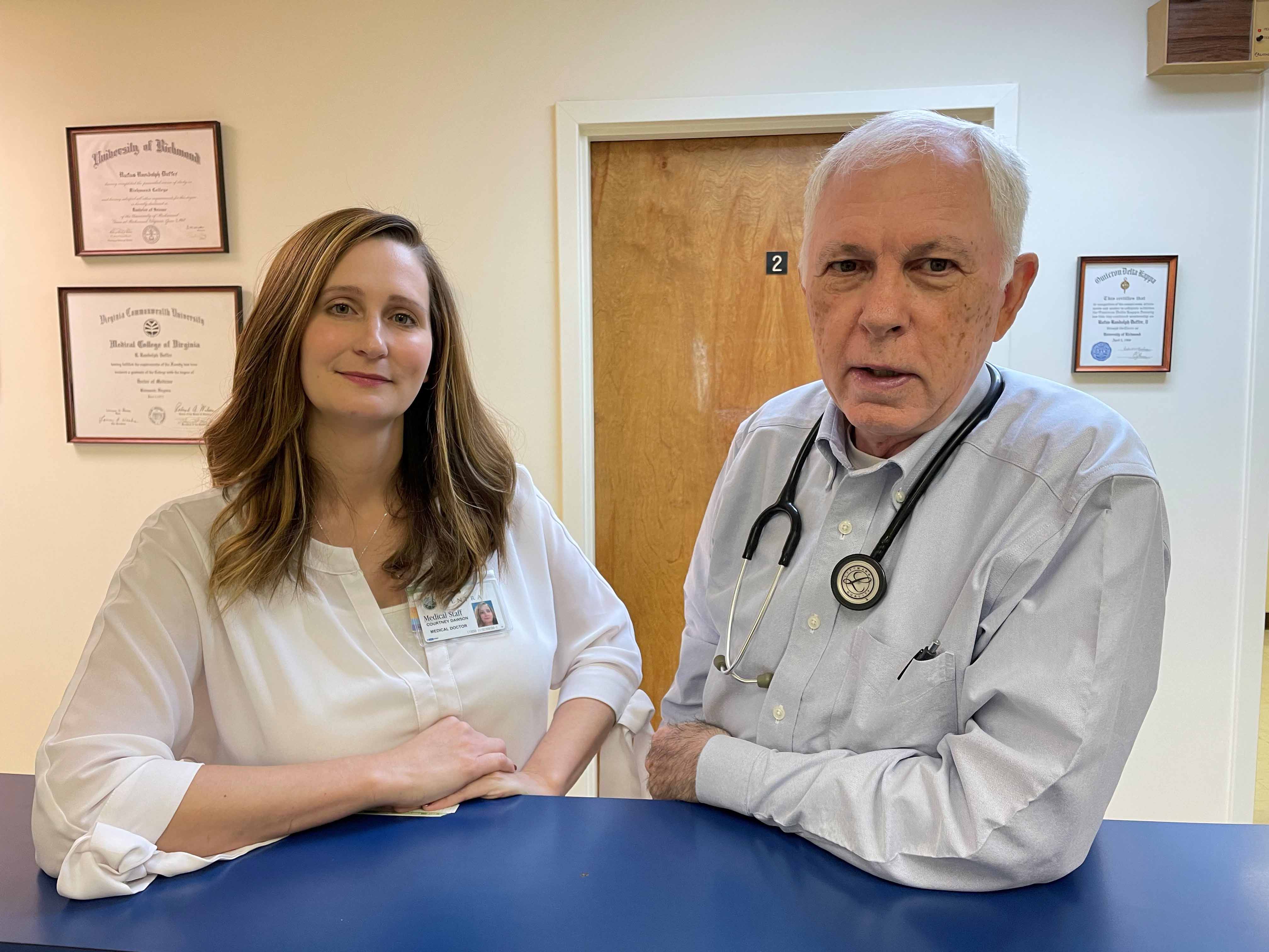 Courtney Dawson, M'18, and Randolph Duffer, M'72, H'75, stand side-by-sde at the reception desk of his rural family practice in Gretna, Virginia.
