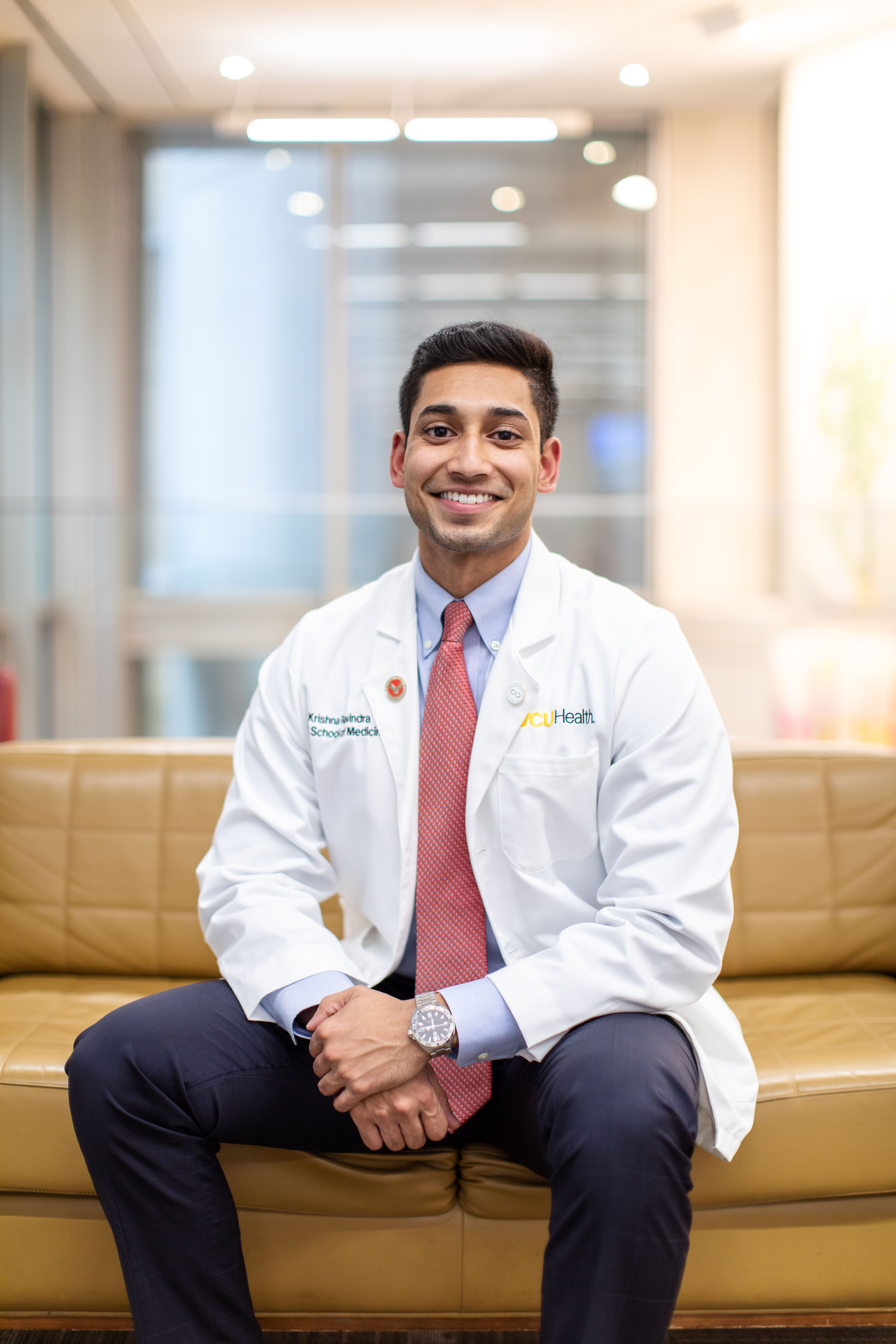 The Class of 2023's Krishna Ravindra smiling in his white coat and sitting on a couch in McGlothlin Medical Education Center