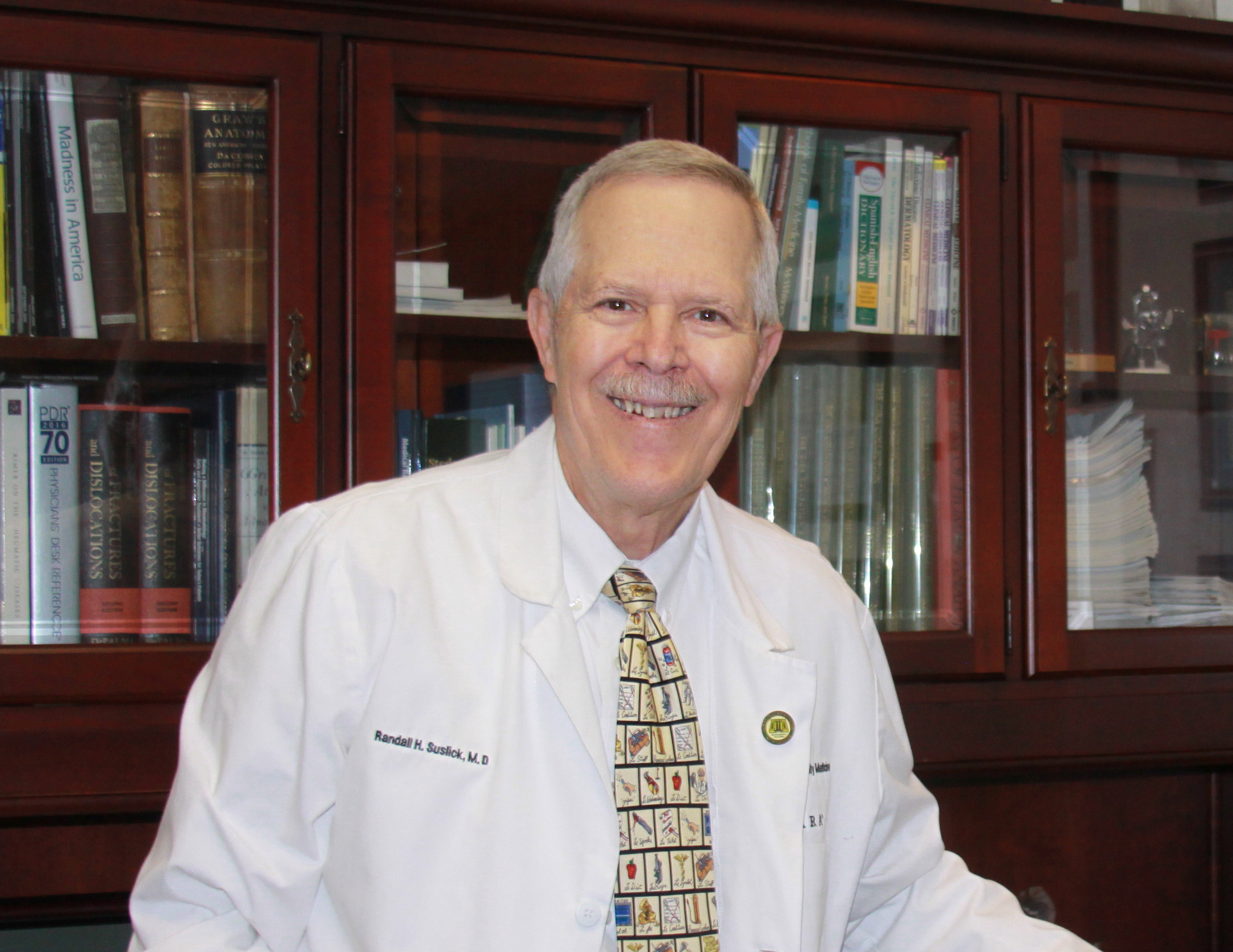 Randall Suslick, M'73, sits at his desk, smiling and wearing his white doctor's coat and a tie